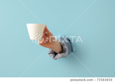 Female hand with cup of tea or coffee breaks through blue paper background. 73563659