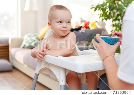 Mother's hand feeds funny cute happy blond baby toddler with spoon Mother's hand feeds funny cute happy blond baby toddler with spoon 73563994