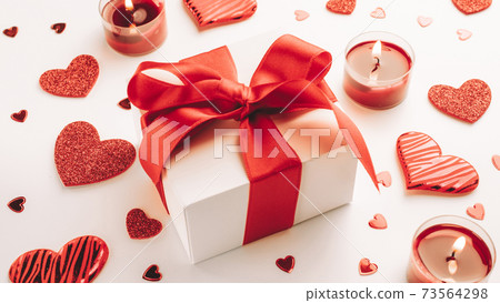 Valentines day background: red love hearts, romantic gift box, candle on white table. February romance present card. Valentines day background: red love hearts, romantic gift box, candle on white table. February romance present card. 73564298