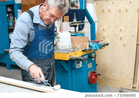An elderly cabinetmaker in overalls and glasses paints a wooden board with a roller on a workbench 73566000