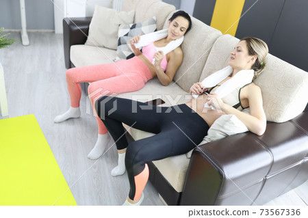 Two tired women are sitting on couch with towel around their necks 73567336