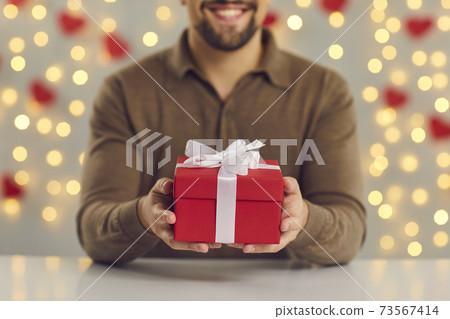 Cropped image close up of a red gift box with a white bow in male hands sitting at the table. Cropped image close up of a red gift box with a white bow in male hands sitting at the table. 73567414
