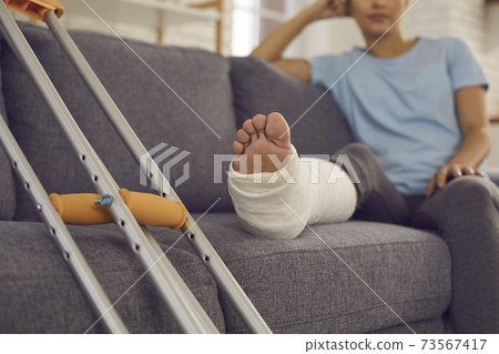 Close up of a crutches and a broken leg in a plaster cast of a woman sitting on a sofa and resting. Close up of a crutches and a broken leg in a plaster cast of a woman sitting on a sofa and resting. 73567417