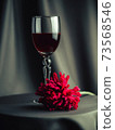 On the table is a glass of red wine. Nearby is a red flower. Artistic blur. 73568546