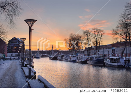 Klaipeda city, Lithuania in January. An old town, river and boats covered by winter snow, cold weather, empty streets. Orange and yellow cloudy sunset sky Klaipeda city, Lithuania in January. An old town, river and boats covered by winter snow, cold weather, empty streets. Orange and yellow cloudy sunset sky 73568611