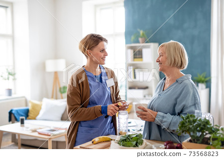 Senior woman with caregiver or healthcare worker indoors, preparing food. 73568823