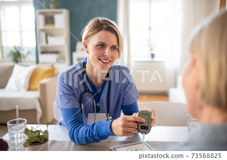 Caregiver or healthcare worker with senior woman patient, measuring blood glucose indoors. Caregiver or healthcare worker with senior woman patient, measuring blood glucose indoors. 73568825