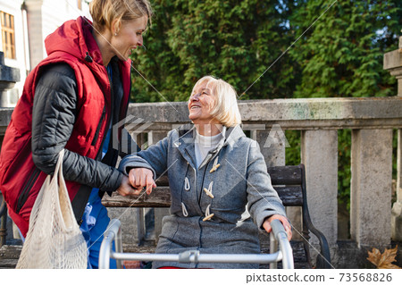Senior woman with walking frame and caregiver outdoors sitting in park, helping to stand up. Senior woman with walking frame and caregiver outdoors sitting in park, helping to stand up. 73568826