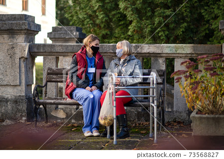 Senior woman with walking frame and caregiver outdoors sitting in park, coronavirus concept. Senior woman with walking frame and caregiver outdoors sitting in park, coronavirus concept. 73568827