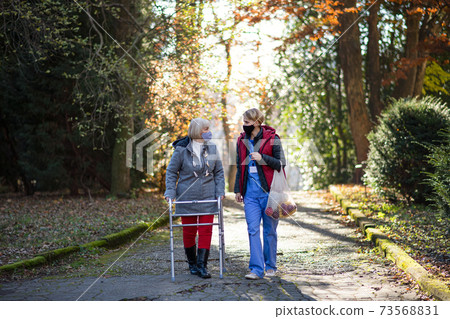 Senior woman with walking frame and caregiver outdoors on a walk in park, coronavirus concept. Senior woman with walking frame and caregiver outdoors on a walk in park, coronavirus concept. 73568831