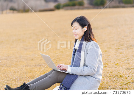 A woman sitting on the grass in the park and typing while listening to music A woman sitting on the grass in the park and typing while listening to music 73573675