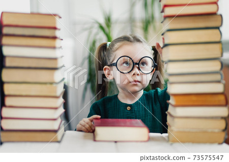 A little beautiful girl with blond hair in round shaped glasses between pile books at home. Stay at home. A little beautiful girl with blond hair in round shaped glasses between pile books at home. Stay at home. 73575547