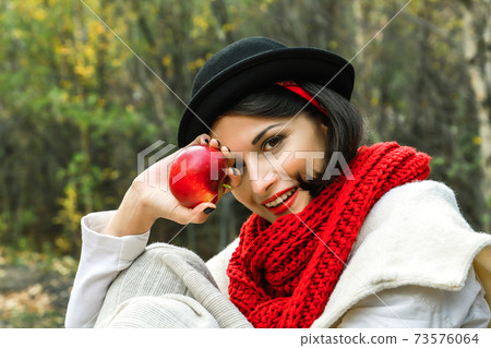 Positive woman in black hat and red scarf with a red apple in her hand smiles and look at the camera. Positive woman in black hat and red scarf with a red apple in her hand smiles and look at the camera. 73576064