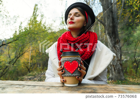 Young woman in a black hat drinks coffee from cozy cup on the nature. Bright smile and good mood Young woman in a black hat drinks coffee from cozy cup on the nature. Bright smile and good mood 73576068