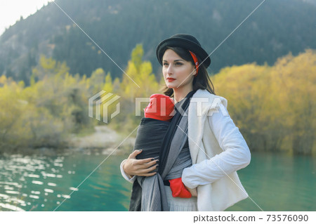 Beautiful young mother with her infant baby in sling outdoor. Mother is carrying her child and travel in autumn mountain lake. Babywearing concept 73576090