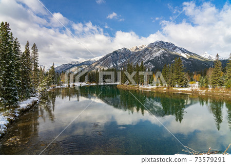 Banff National Park beautiful natural scenery in winter sunny day. Mount Norquay with blue sky, white clouds reflected on Bow River like a mirror. Town of Banff, Canadian Rockies. Banff National Park beautiful natural scenery in winter sunny day. Mount Norquay with blue sky, white clouds reflected on Bow River like a mirror. Town of Banff, Canadian Rockies. 73579291