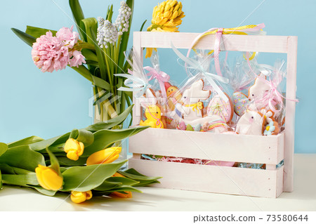 Easter cookies in personal package bag with pastel colored icing in wooden box and spring flowers on blue background. Happy easter holiday card, set of sweet gifts for anniversary, cute gingerbread Easter cookies in personal package bag with pastel colored icing in wooden box and spring flowers on blue background. Happy easter holiday card, set of sweet gifts for anniversary, cute gingerbread 73580644