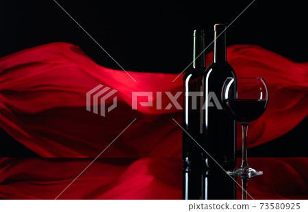 Wineglass and bottles of red wine on a black reflective background. Red satin curtain flutters in the wind. Wineglass and bottles of red wine on a black reflective background. Red satin curtain flutters in the wind. 73580925