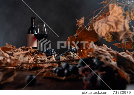 Bottle and glass of red wine on a table with dried vine leaves and blue grapes. 73580979