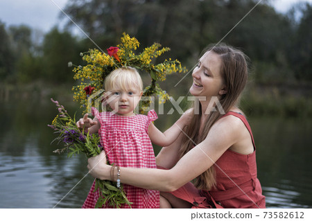 Mom and little daughter are resting by the river. Child in wreath of flowers. 73582612