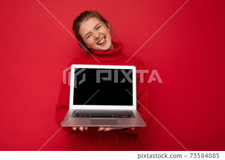 Beautiful satisfied happy young woman holding computer laptop looking at camera wearing red sweater 73584085
