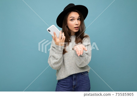 Beautiful young asking dissatisfied brunette woman wearing black hat and grey sweater holding 73585905