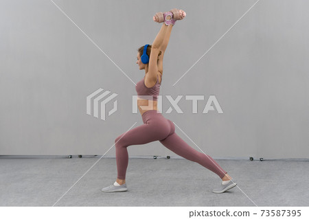 Stretching using dumbbells lifted hands up young girl losing weight at home wearing pink outfit while listen music in blue wireless headphones. Sport and recreation concept Stretching using dumbbells lifted hands up young girl losing weight at home wearing pink outfit while listen music in blue wireless headphones. Sport and recreation concept 73587395
