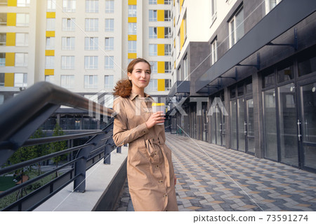 Portrait of a young successful woman in beige trech looking into the distance on a background of high-rise building 73591274
