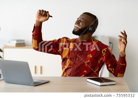 Joyful african freelancer man having fun at workplace, singing and listening music Joyful african freelancer man having fun at workplace, singing and listening music 73595332