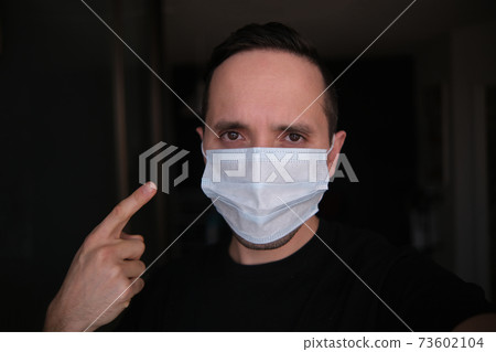 A young man puts on a protective medical mask, a close-up of a European guy removing a surgical mask during the Covid-19 pandemic on a dark blurry background A young man puts on a protective medical mask, a close-up of a European guy removing a surgical mask during the Covid-19 pandemic on a dark blurry background 73602104