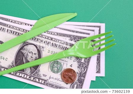 Disposable plastic cutlery green. Plastic fork and knife lie on a small amount of US dollars Disposable plastic cutlery green. Plastic fork and knife lie on a small amount of US dollars 73602139