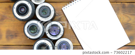 Several photographic lenses and white notebook lie on a brown wooden background. Space for text 73602217