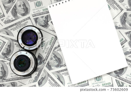 Two photographic lenses and white notebook lie on the background of a lot of dollar bills. Space for text Two photographic lenses and white notebook lie on the background of a lot of dollar bills. Space for text 73602609
