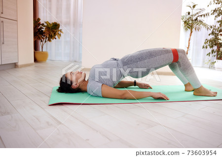 Young athlete performs a glute bridge on a fitness mat using an elastic band 73603954