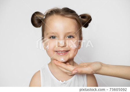 Mother hand applying cream on face of pretty little smiling 3 years old girl after bathing, isolated on white background. Close up portrait. Promo ad of children's cosmetics 73605092