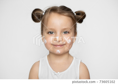 Close up of funny cheerful little 3 years old blond haired Caucasian girl in white shirt with cream for face skin, isolated on white background. Spa skin care treatments, baby skin protection Close up of funny cheerful little 3 years old blond haired Caucasian girl in white shirt with cream for face skin, isolated on white background. Spa skin care treatments, baby skin protection 73605102
