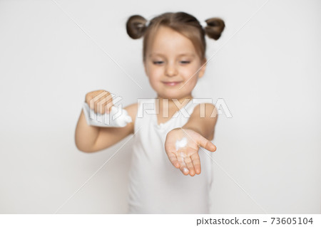 Cheerful cute little girl holding tube with protective cream, and showing her little hand with cream to camera. Close up portrait on isolated white background. Focus on the hand Cheerful cute little girl holding tube with protective cream, and showing her little hand with cream to camera. Close up portrait on isolated white background. Focus on the hand 73605104