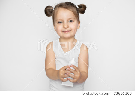 A little girl with blonde hair in two ponytails, wearing white top, holding tube of cream. Promo of children's cosmetics, skin care products. Isolated on white background A little girl with blonde hair in two ponytails, wearing white top, holding tube of cream. Promo of children's cosmetics, skin care products. Isolated on white background 73605106