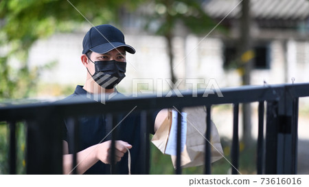 Deliver man in protective mask  holding paper bag with food and waiting customer at doorway. Food delivery service. 73616016