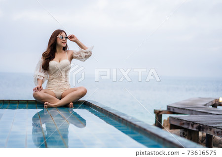 woman sitting on edge of swimming pool and looking to the sea woman sitting on edge of swimming pool and looking to the sea 73616575