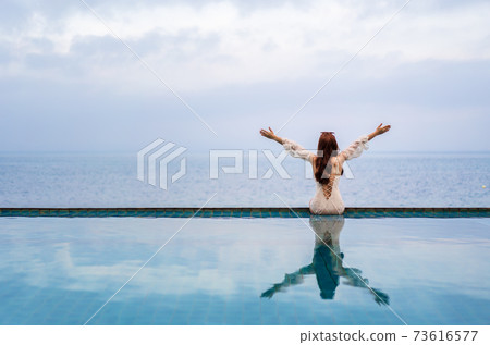 woman sitting with arms raised on edge of swimming pool and looking to the sea woman sitting with arms raised on edge of swimming pool and looking to the sea 73616577