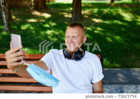 Man in medical mask sitting on the bench in the park take selfie by mobile phone. Man in medical mask sitting on the bench in the park take selfie by mobile phone. 73625754