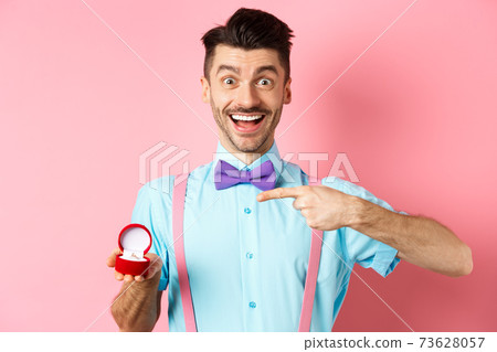 Valentines day. Cheerful young man going to make proposal, pointing finger at engagement ring and smiling excited, standing on romantic pink background 73628057