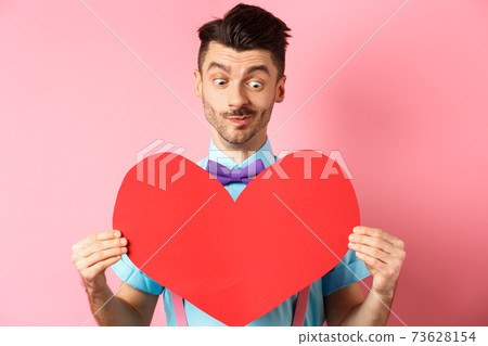 Valentines day concept. Cute boyfriend looking at big red heart, waiting for lover on romantic date, pink background Valentines day concept. Cute boyfriend looking at big red heart, waiting for lover on romantic date, pink background 73628154