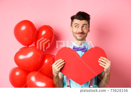 Valentines day concept. Smiling handsome man looking dreamy left, waiting for true love girlfriend with big red heart cutout, standing over pink background Valentines day concept. Smiling handsome man looking dreamy left, waiting for true love girlfriend with big red heart cutout, standing over pink background 73628156