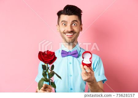 Valentines day. Smiling handsome man asking to marry him, showing engagement ring and red rose, standing romantic on pink background 73628157