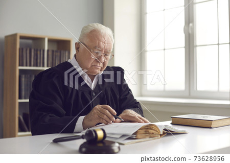 Serious wise mature judge in gown sitting at desk in courtroom and reading a book Serious wise mature judge in gown sitting at desk in courtroom and reading a book 73628956