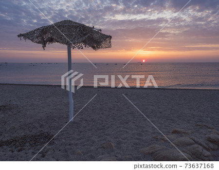 Colorful sunrise at empty beach Spiaggia di Santa Maria Navarrese with wooden parasol, red sun raising up from the sea and pink blue and orange clouds. Sardinia, Italy Colorful sunrise at empty beach Spiaggia di Santa Maria Navarrese with wooden parasol, red sun raising up from the sea and pink blue and orange clouds. Sardinia, Italy 73637168