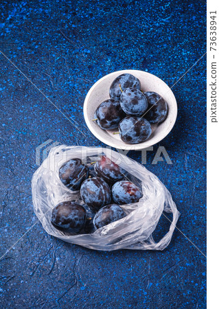 Fresh ripe plum fruits in plastic bag package and in wooden bowl on blue abstract background, angle view 73638941