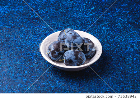 Fresh ripe plum fruits in white wooden bowl on blue abstract background, angle view Fresh ripe plum fruits in white wooden bowl on blue abstract background, angle view 73638942